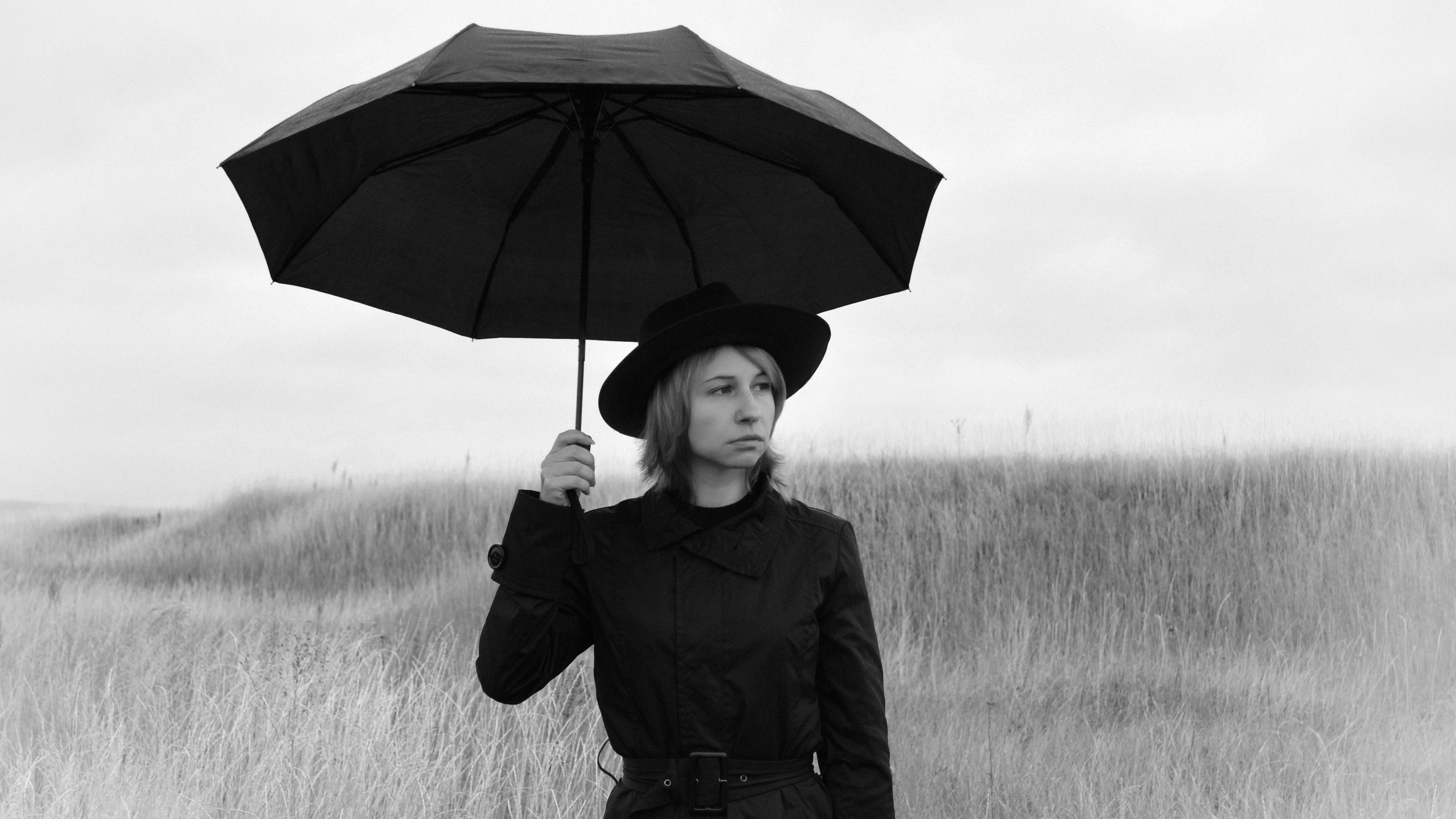 Serious young woman in coat and hat holding umbrella in gloomy field, capturing a psychological thriller mood
