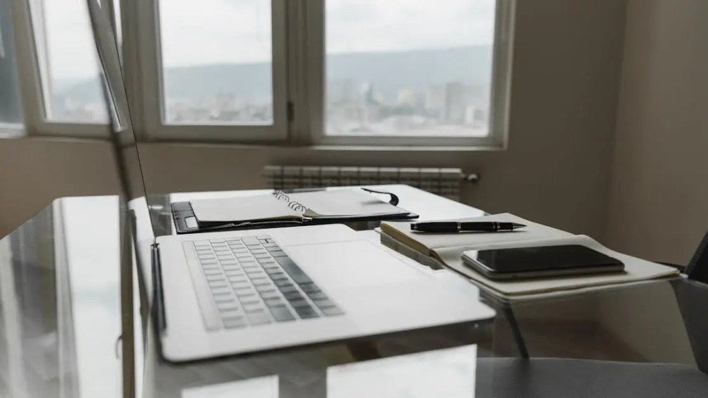 Laptop, open notebook, pen, and smartphone on a glass desk in a modern workspace, set up for pitching a screenplay.