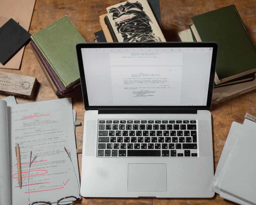 An overhead view of a laptop displaying a screenplay document, surrounded by stacks of books, loose printed pages covered in red editorial markups, a pen, and glasses on a wooden desk.