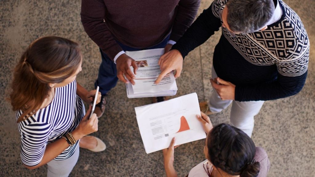 Film producers and executives standing in a circle reviewing script coverage reports and financial projection charts for film financing decisions.