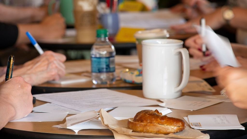 A group of screenwriters gathered around a table with notebooks, scripts, and coffee cups during a collaborative writers' room brainstorm session.