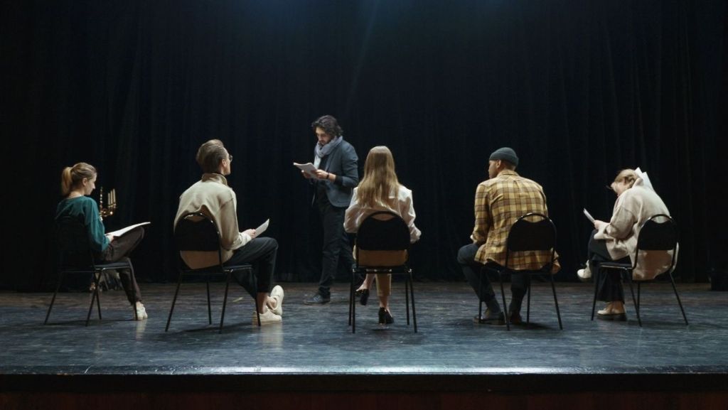 Ensemble cast of actors seated in a semi-circle on stage reading scripts, representing multiple protagonists and balanced character arcs.